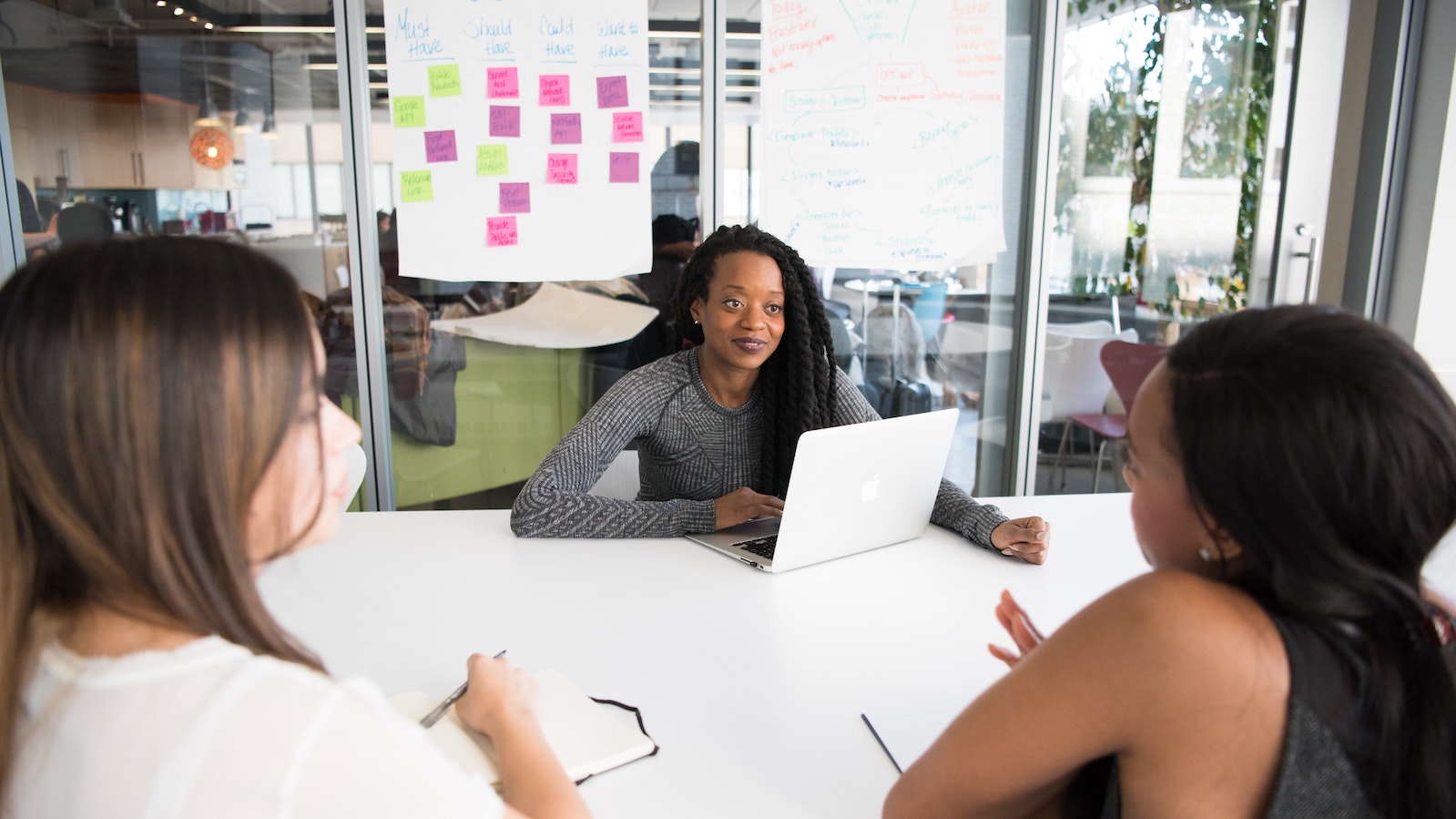 Two women at a work table facing another woman who has a laptop