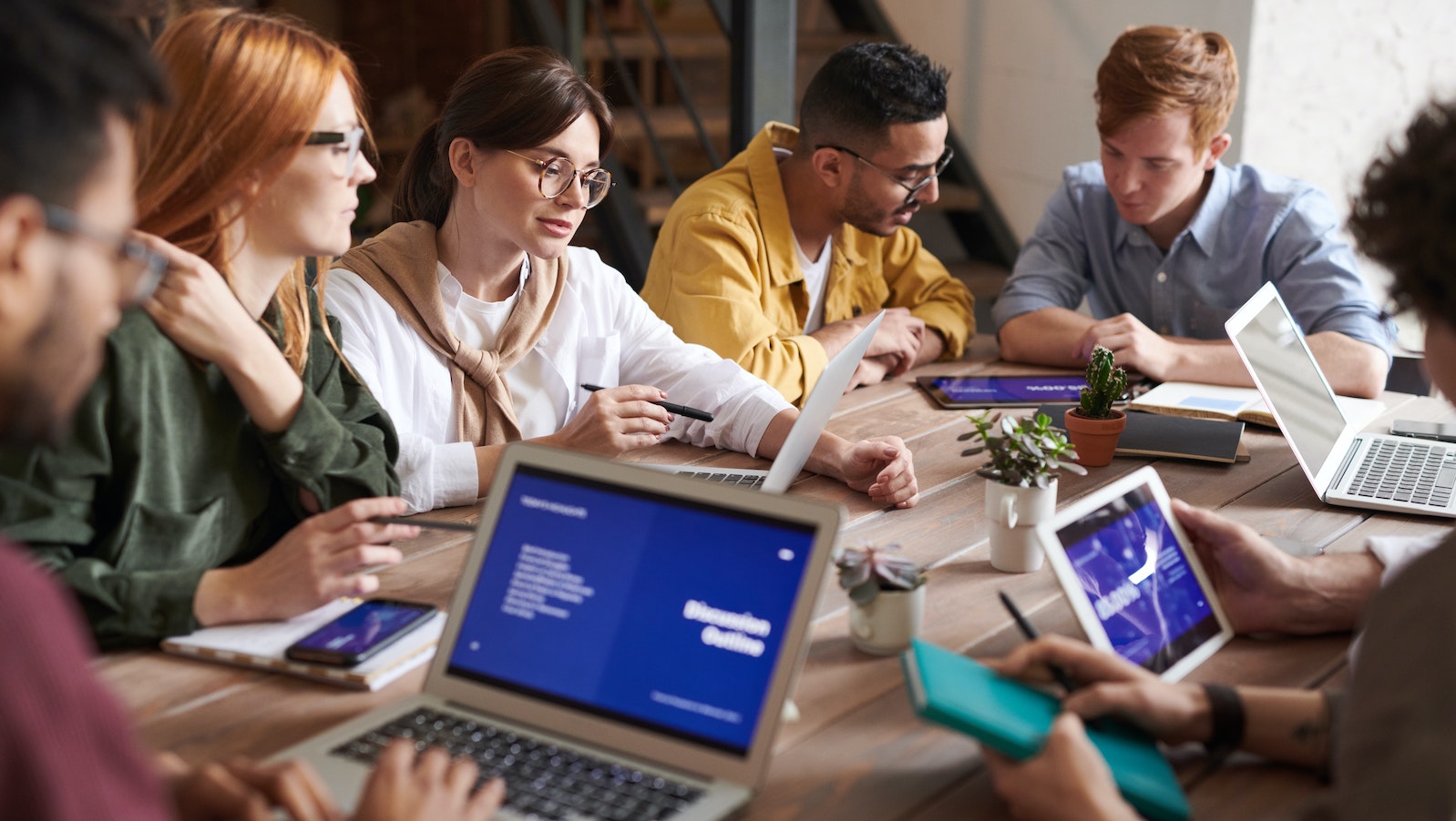 A group of mid-20s people sitting around a desk talking and with laptops open