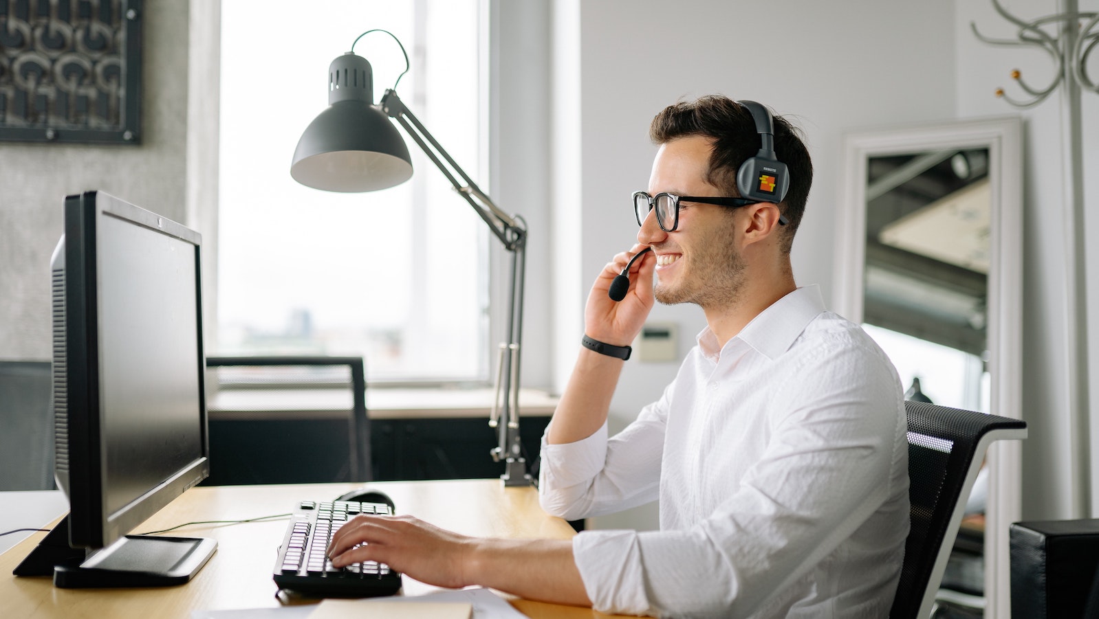 A man smiling wearing a headset sitting at a desk and looking at a computer