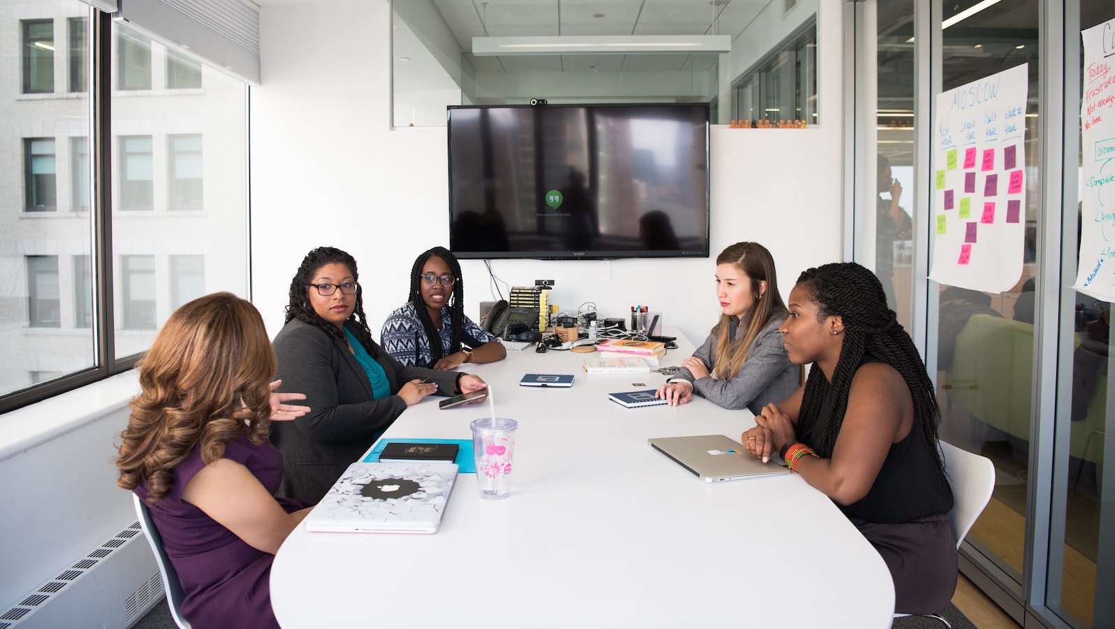 Five women sitting at a white desk in an office talking with computers in front of them