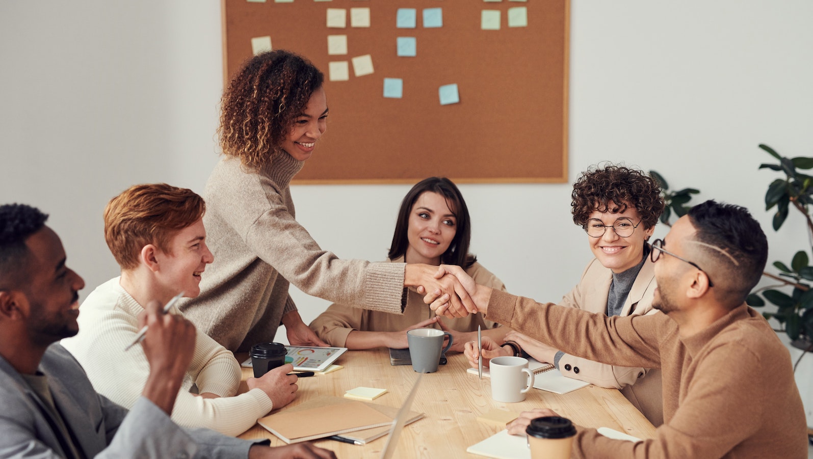 Six people around a desk with a bulletin board in the back. Two people are shaking hands across the table