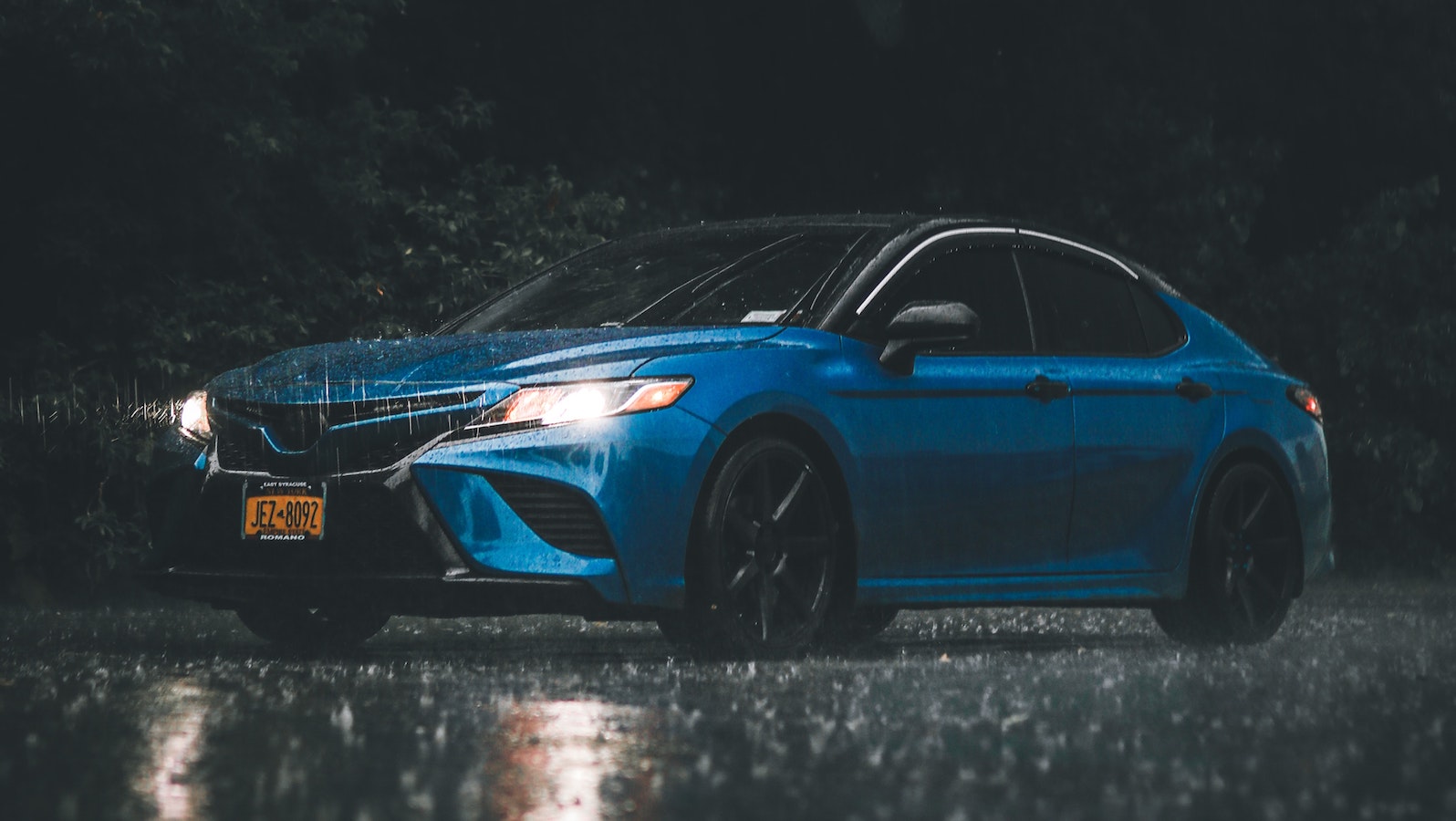 A blue car on a road at night with lights on while it's raining