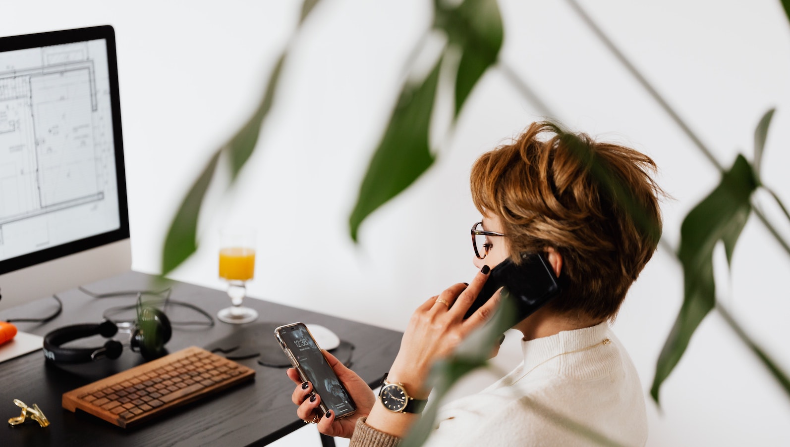 A woman sitting at her desk on the phone while holding another phone and in front of a desktop computer