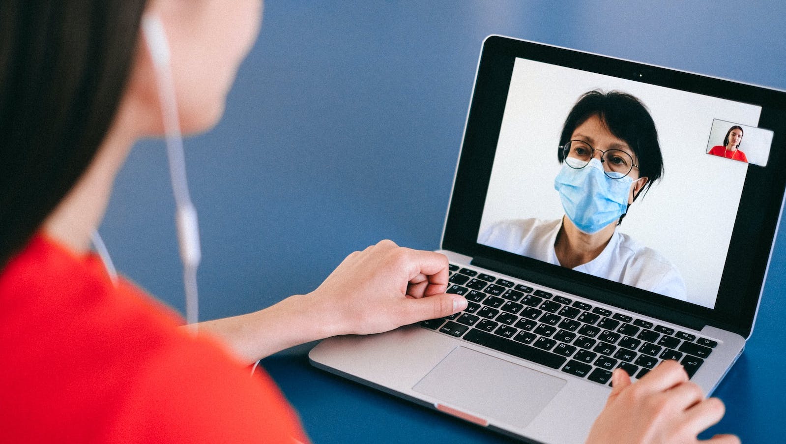 A woman with headphones on talking to a doctor wearing a mask via laptop