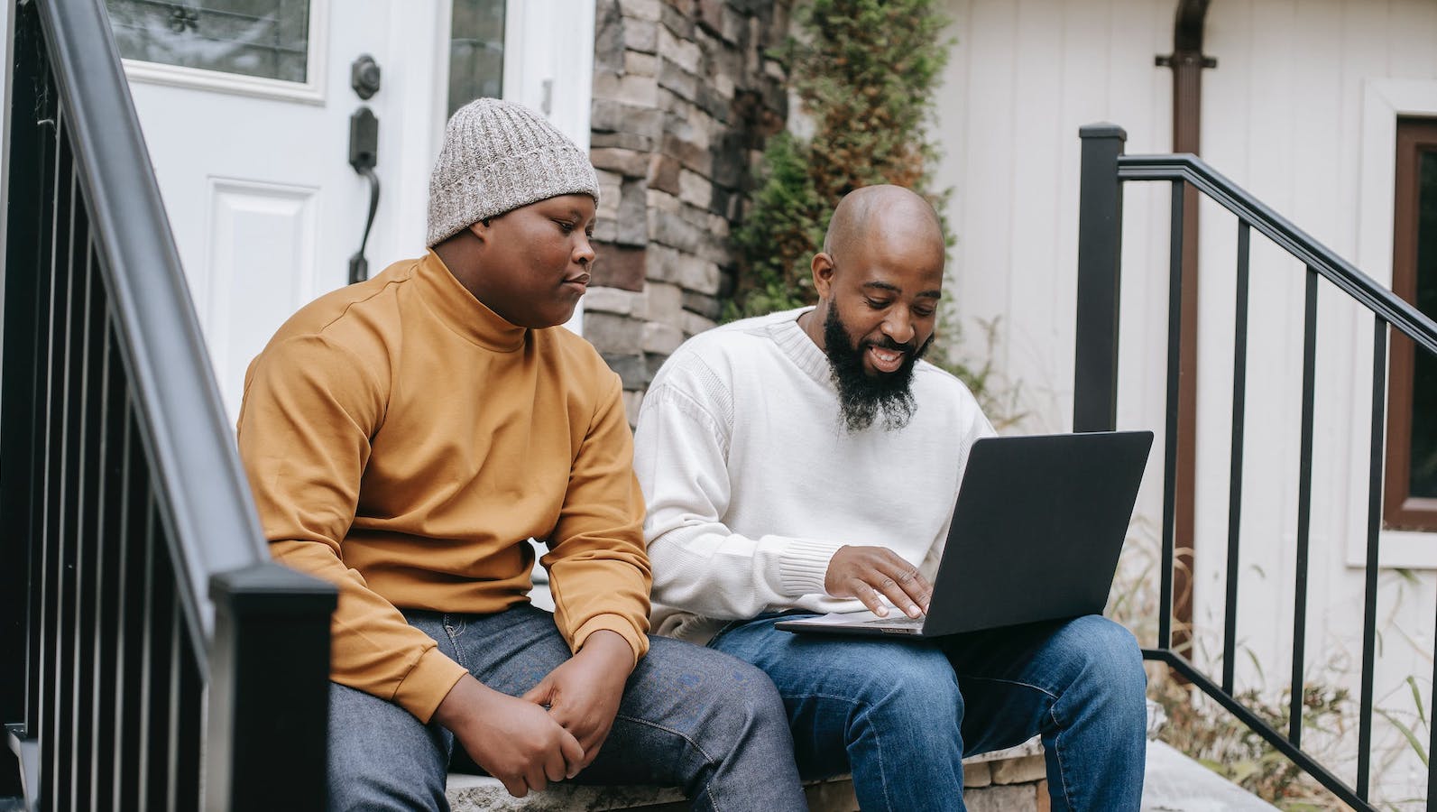Two men sitting on front door steps side by side with a black laptop