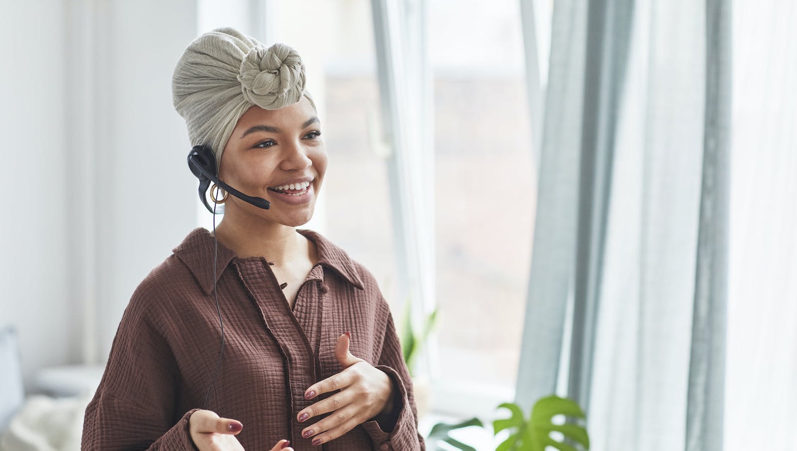 A woman smiling while wearing a brown top and a head scarf standing in front of a window with a black headset on