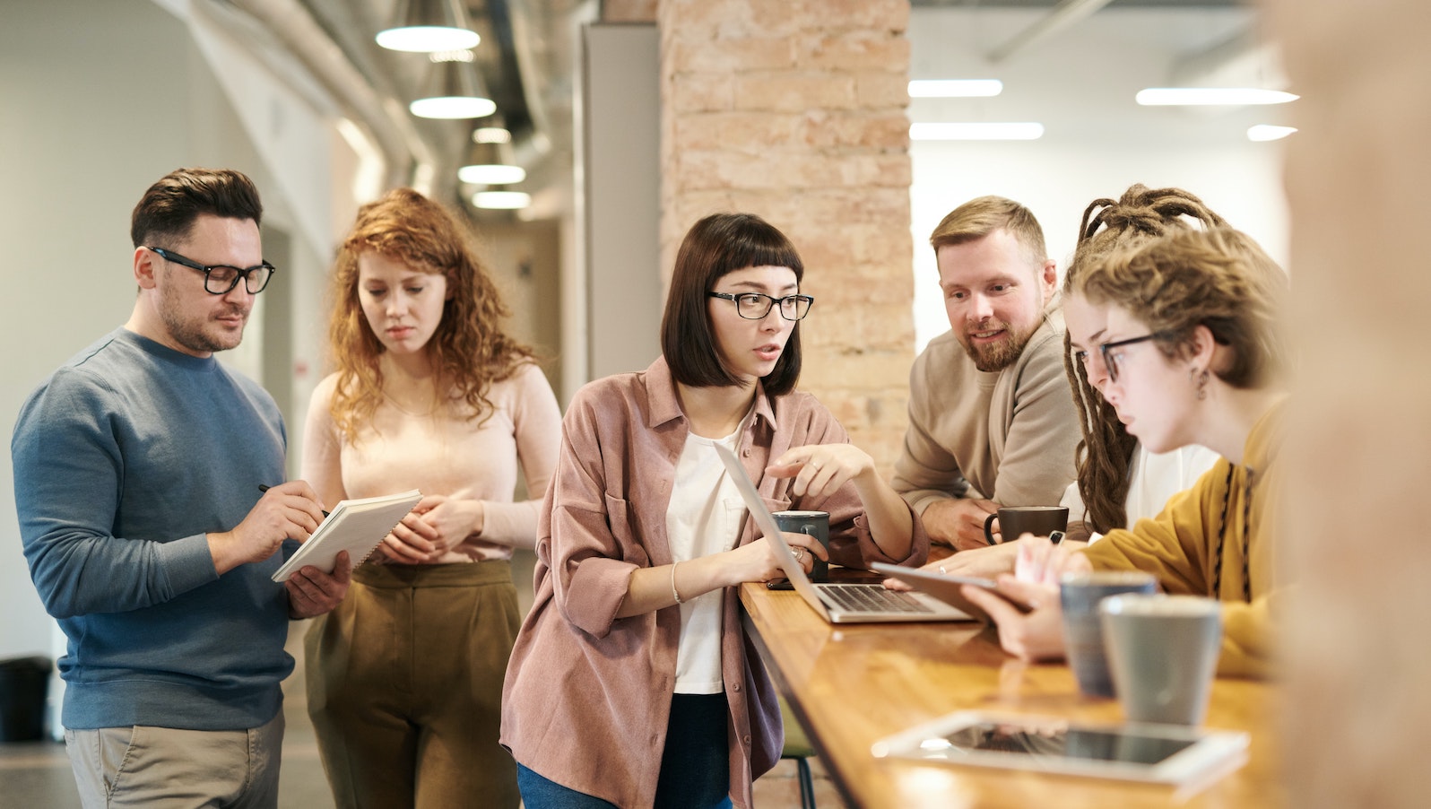 A group of people standing around tablets at an office table while talking and drinking coffee in mugs