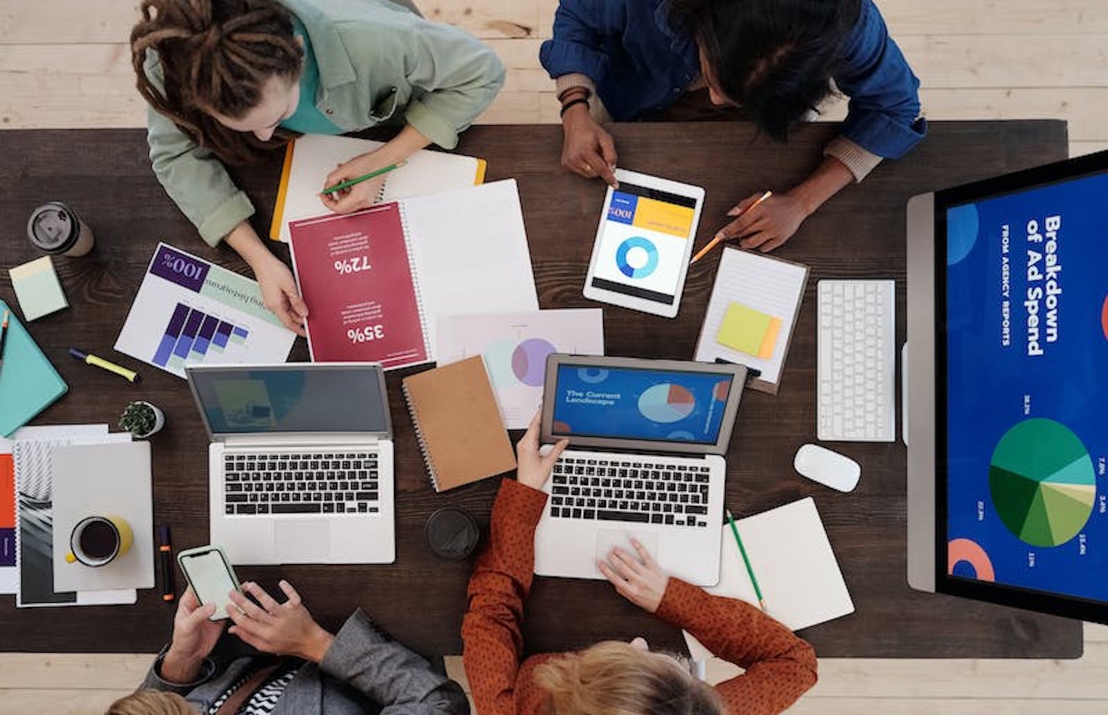 Overhead view of four people at a brown desk with laptops and charts in front of them