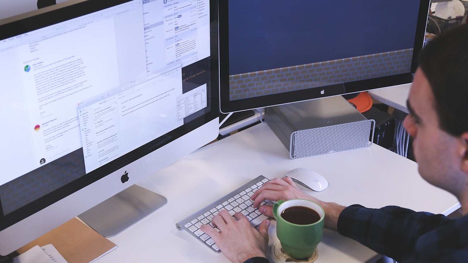 Man at a desk with a computer open and typing on a keyboard with a green mug of coffee in front of him