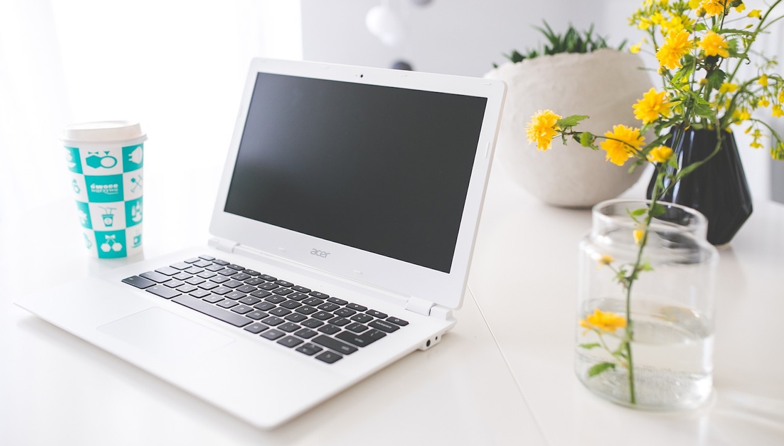 Laptop on a white counter with yellow flowers in a glass next to it and a blue glass