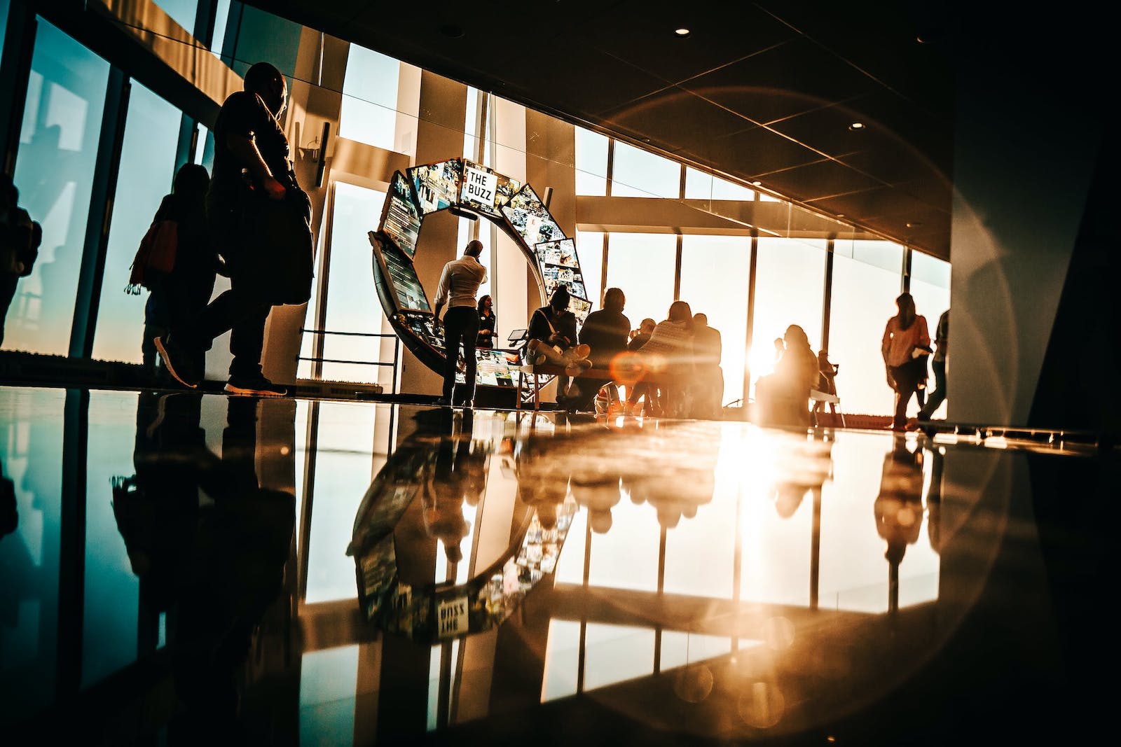 A group of people standing in front of a screen in a building with glass windows and sunlight coming through