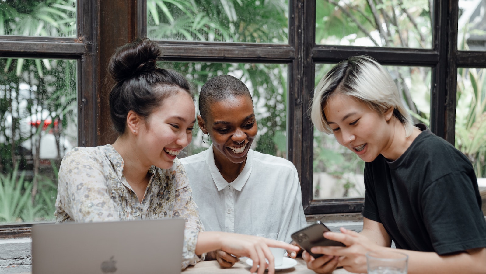 Three women sitting at a table with a laptop in front of them and looking and smiling at a phone