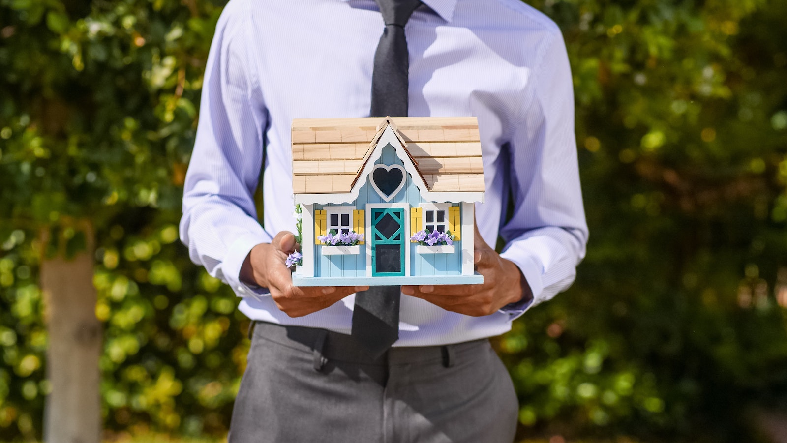 Person in a suit holding a miniature house against a background of trees