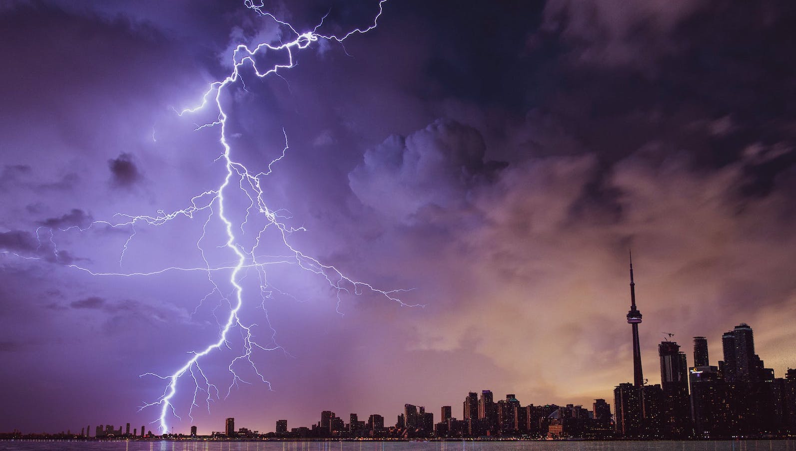 A dark city skyline with clouds overhead and a large bolt of lighting against a purple sky