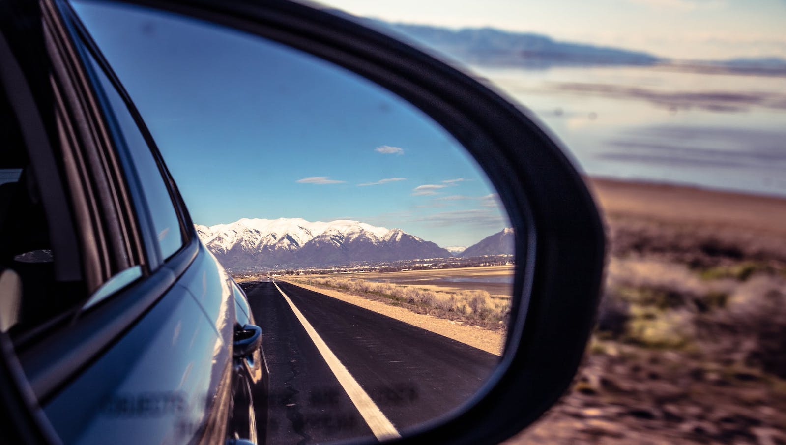 Close-up image of a car's side mirror showing a reflection of the road and mountains and blue sky behind it alongside an image in the foreground