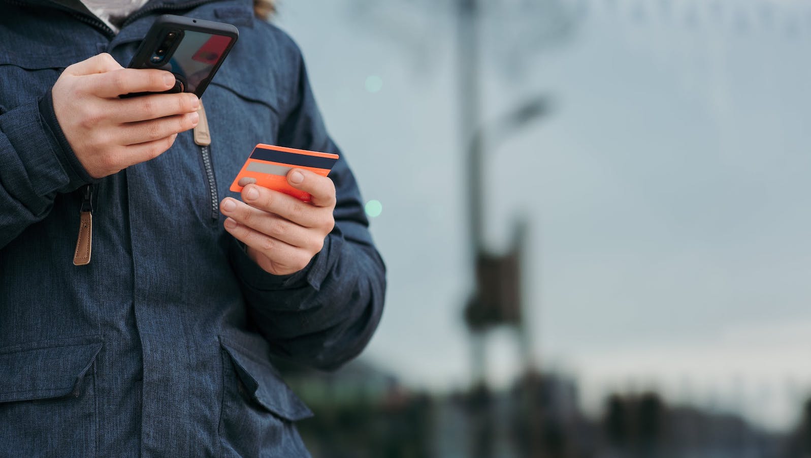 Person in a blue jacket holding a phone and a red credit card set against a blurry background