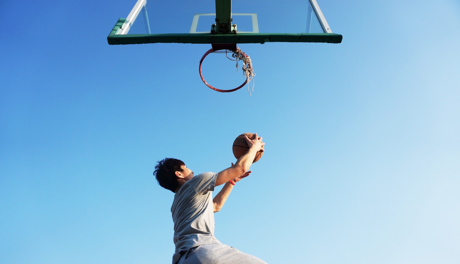 Low angle shot of a person with a basketball going for a layup on a basketball hoop against a light blue sky