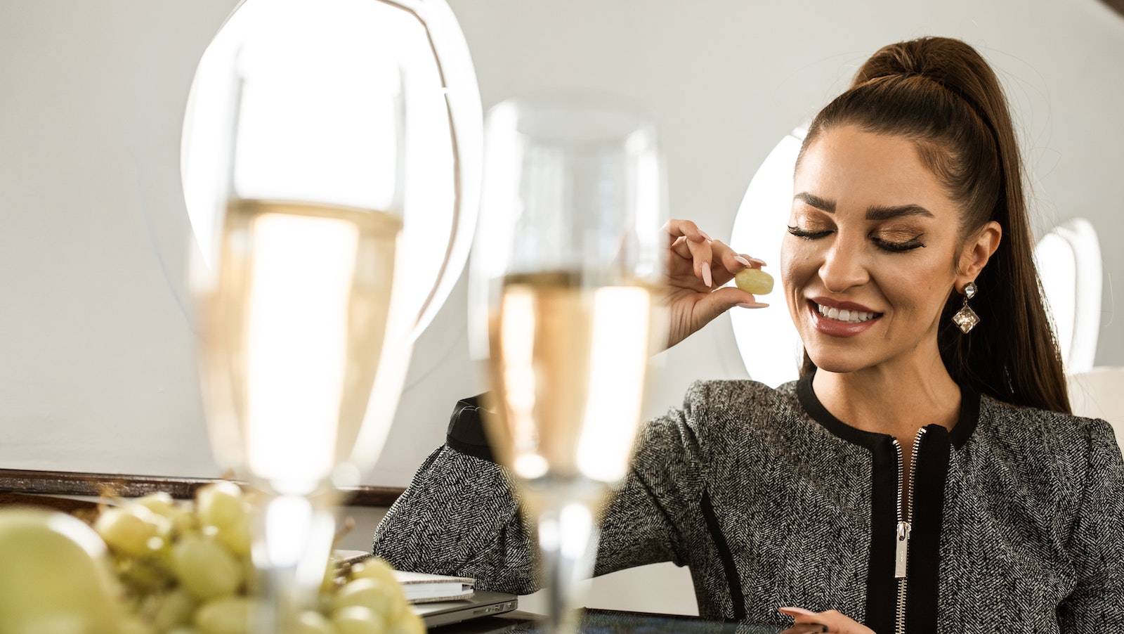 Woman sitting in the window seat on an airplane wearing a suit while in front of two glasses of champagne and grapes