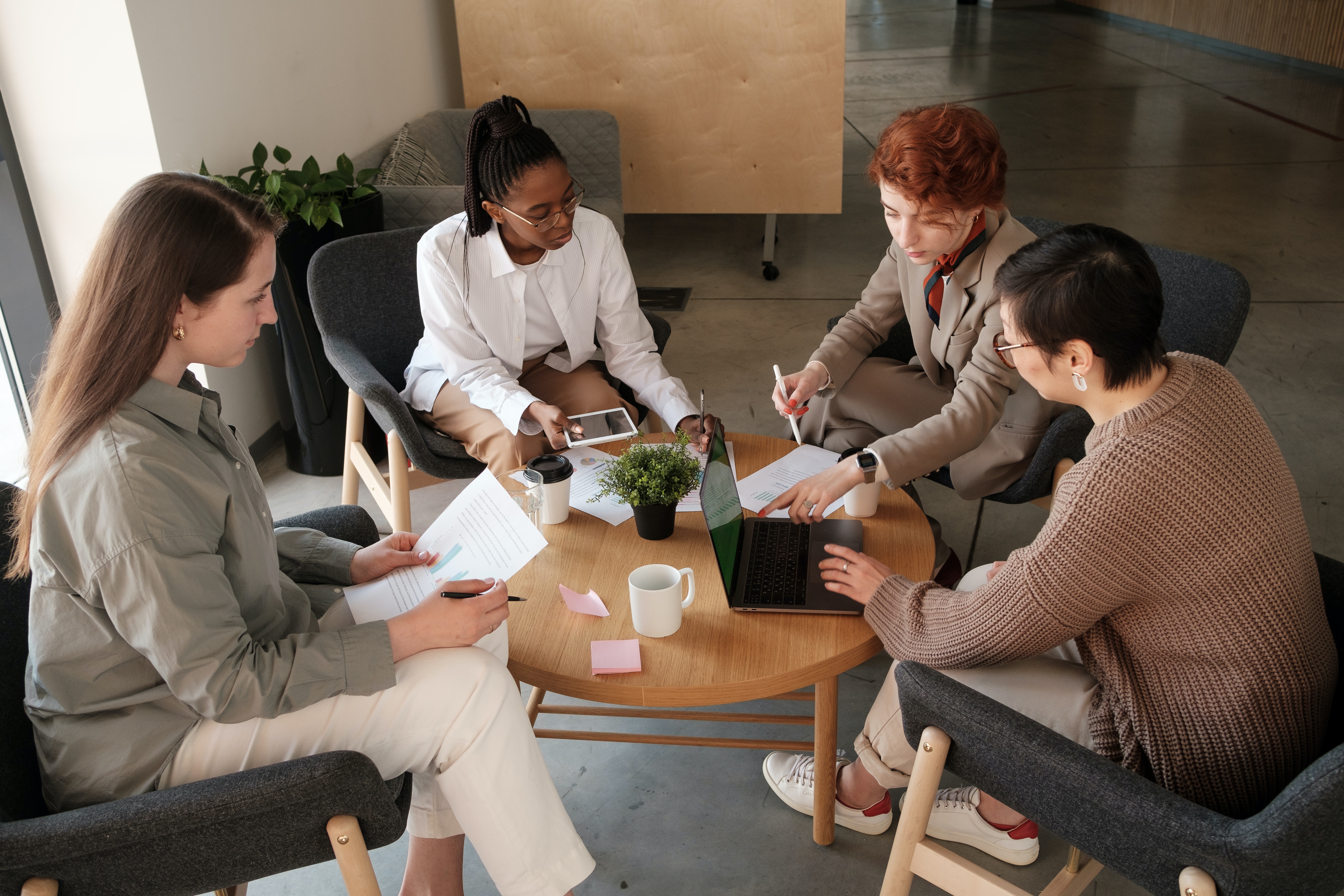 Four women sitting around a brown wooden circular table with notes and tablets in front of them