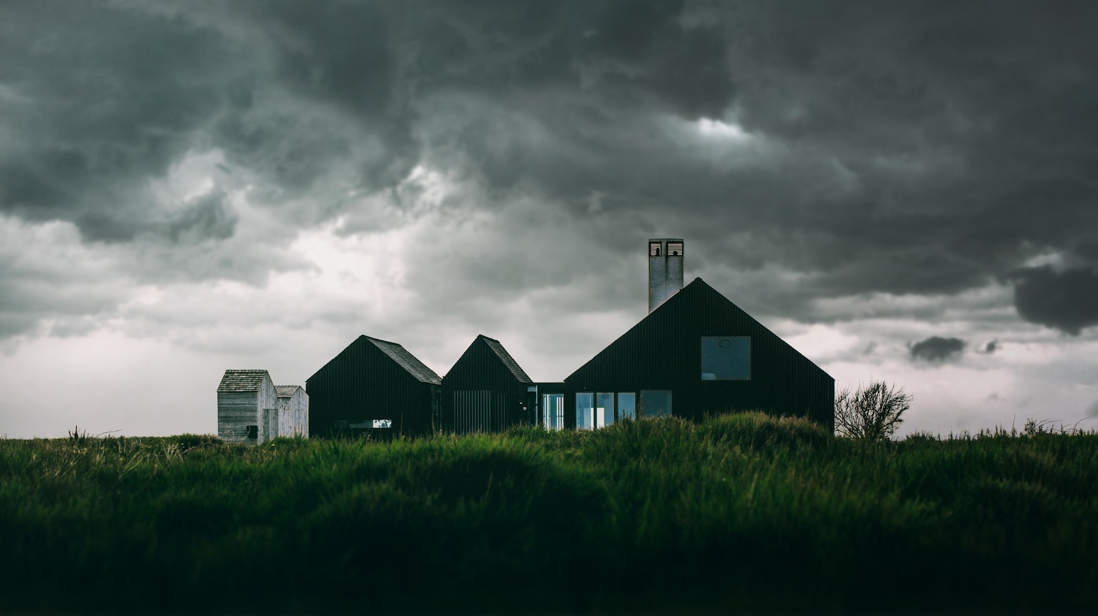 A simple brown house in the countryside atop of tall green grass and beneath a dark and cloudy sky