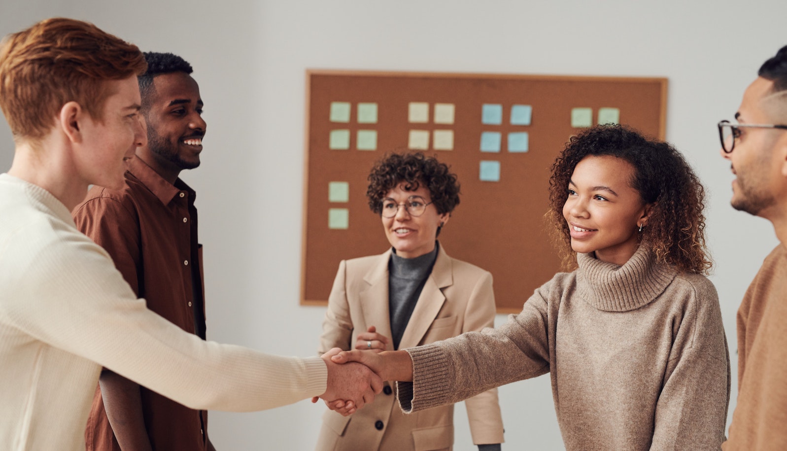 Give people standing and all smiling while two people shake hands with a bulletin board with post-it notes in the background