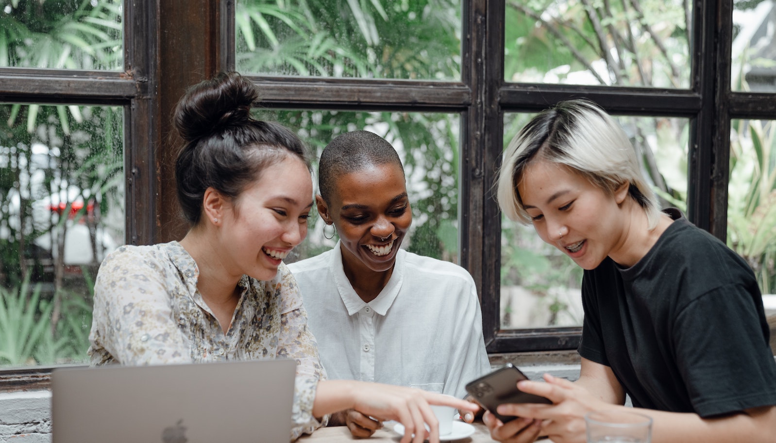 Three young women sitting at a table looking at an electronic device and smiling