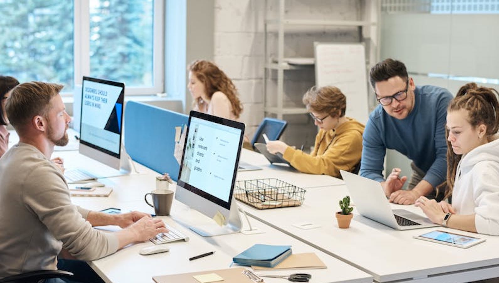 A multitude of people in an office sitting at a desk and looking at their computers and talking to one another