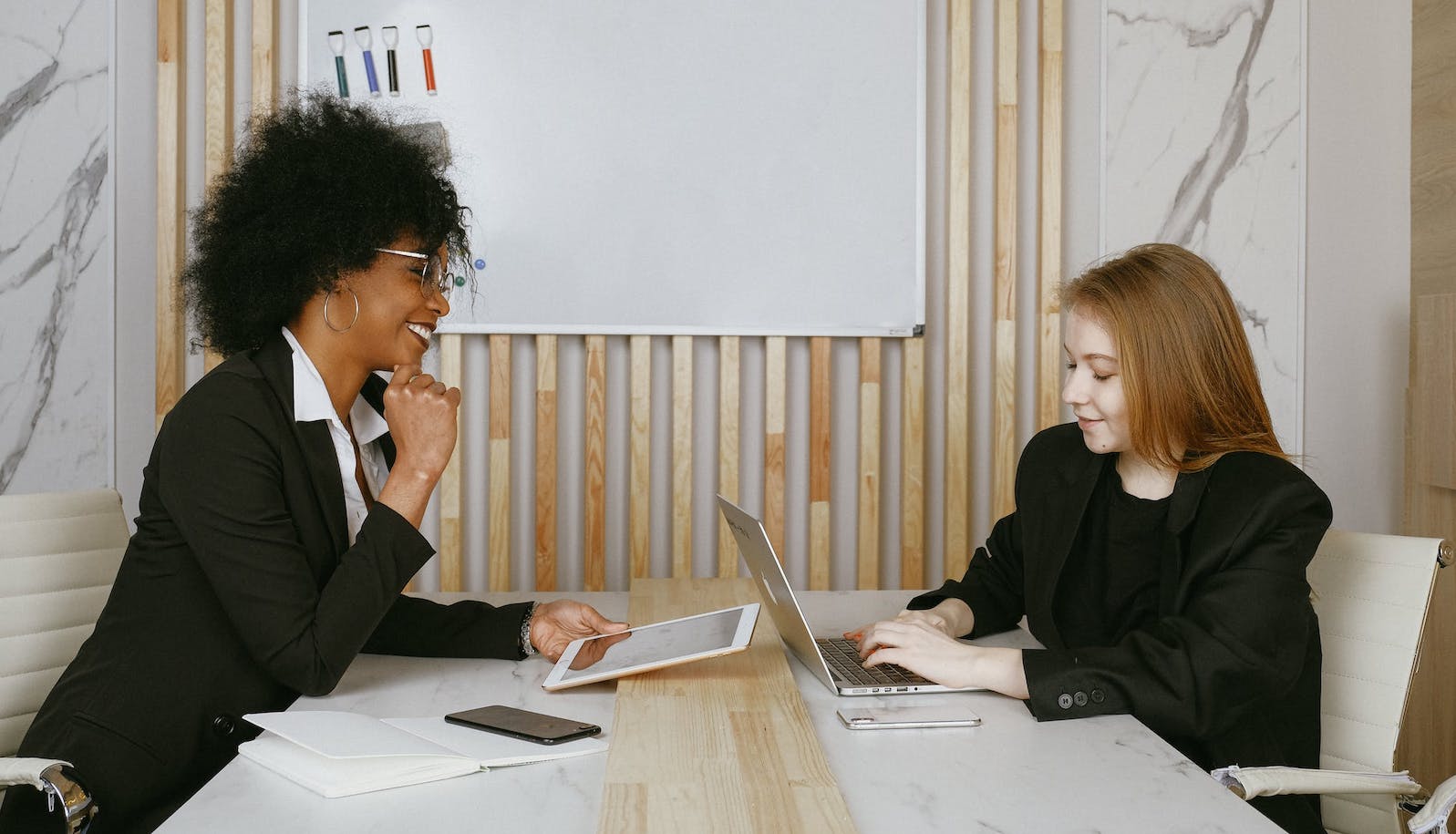 Two women sitting across from each other at a table and smiling and looking at their laptops