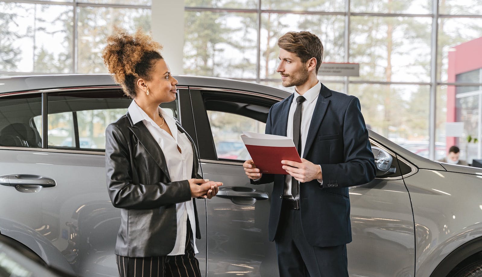 Two people at a car dealership in front of a car and talking to each other