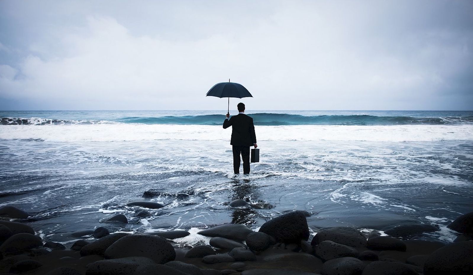 A man in a suit holding a briefcase and an umbrella standing in the ocean under a cloudy sky