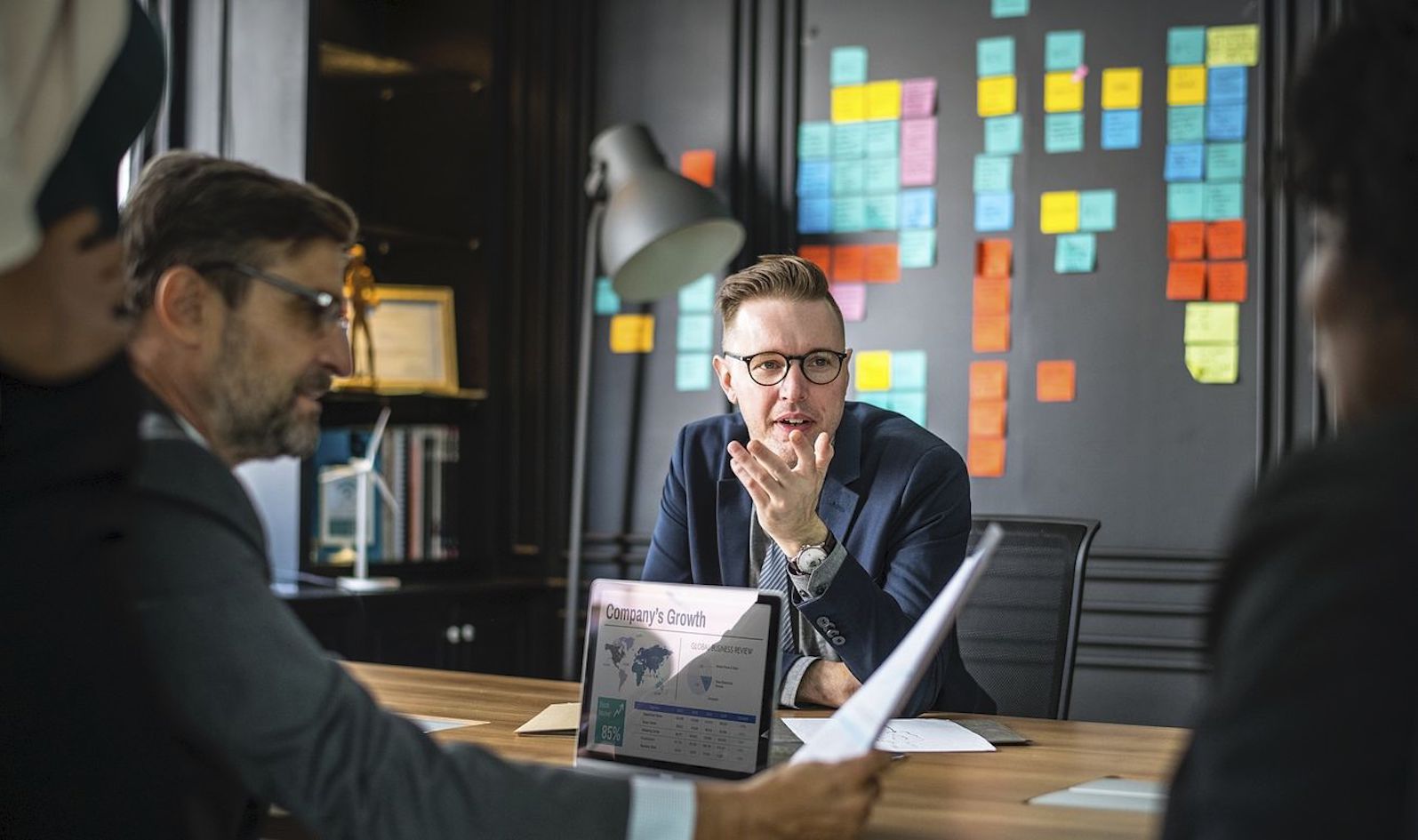 Three people sitting at a table and one person standing, and they're all talking; there are papers and a laptop on the table and behind the people is a black wall with post-it notes covering it