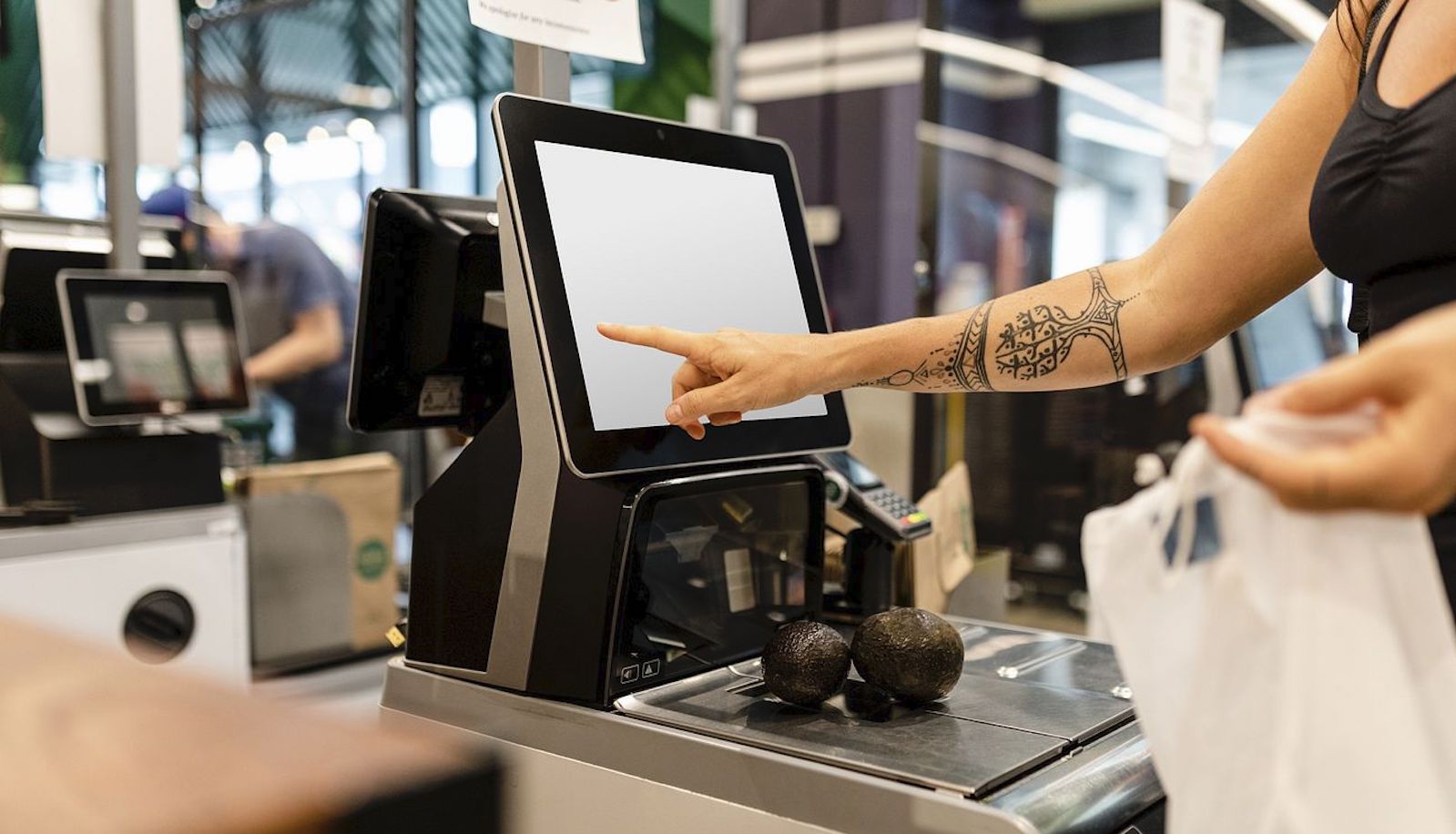 A woman touching the screen at a self checkout stand in a store with two avocados sitting on the scale