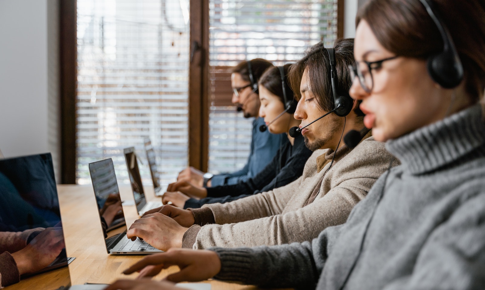 Four people in a row sitting at a desk in front of laptops with headsets on