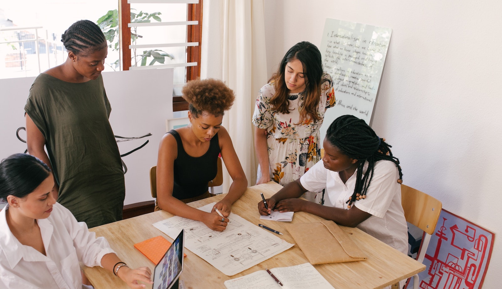 Five women sitting and standing at a desk while working on papers and also on laptops