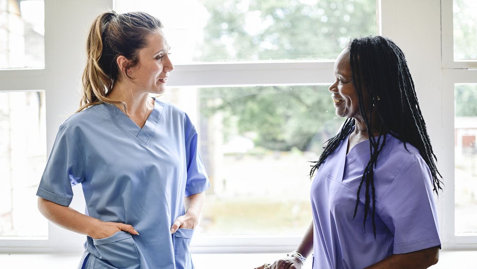 Two women in blue and purple scrubs talking to each other in a hospital