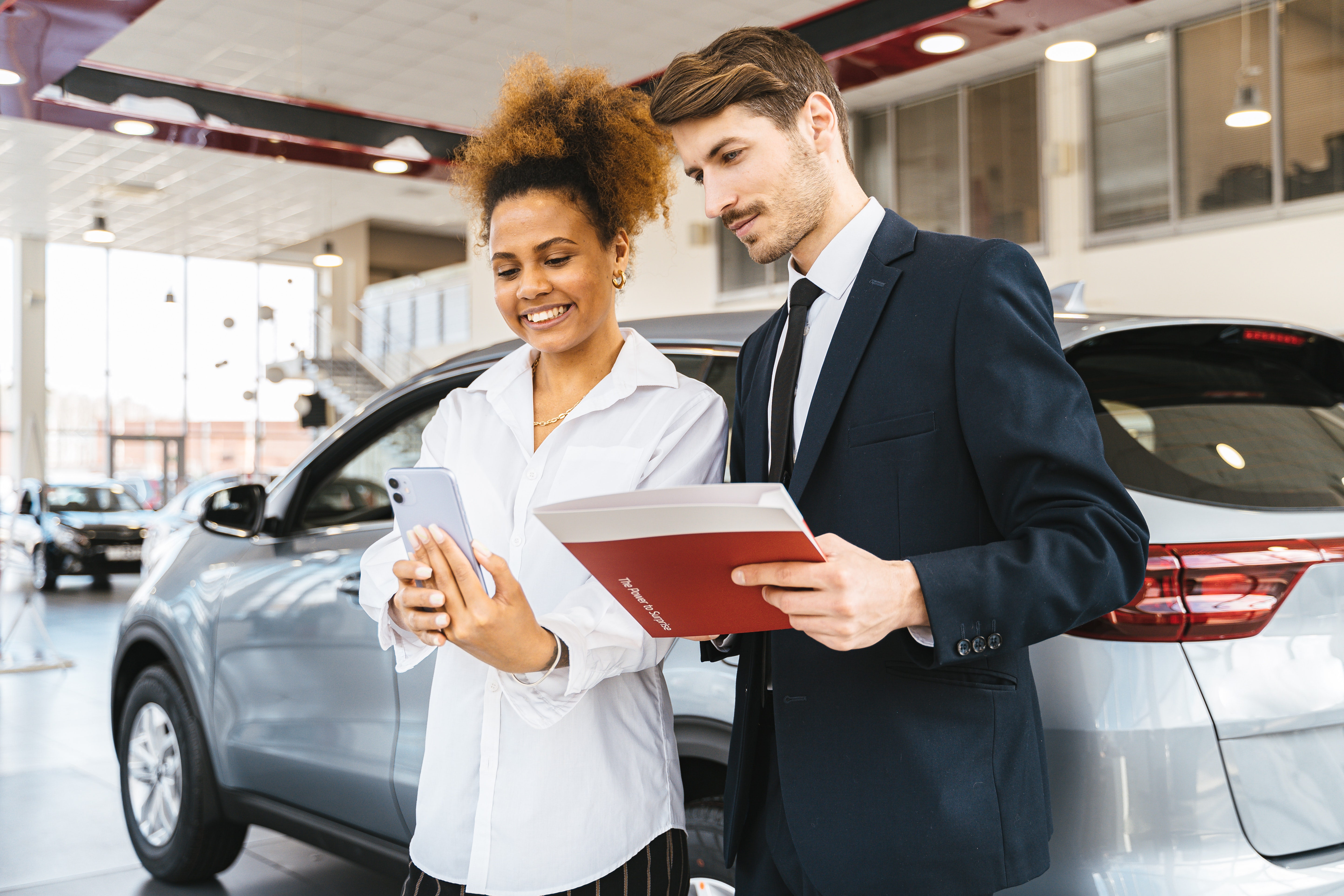 A man and a woman in front of a silver mid-size car in a car dealership while they're both looking at her phone and he's holding papers