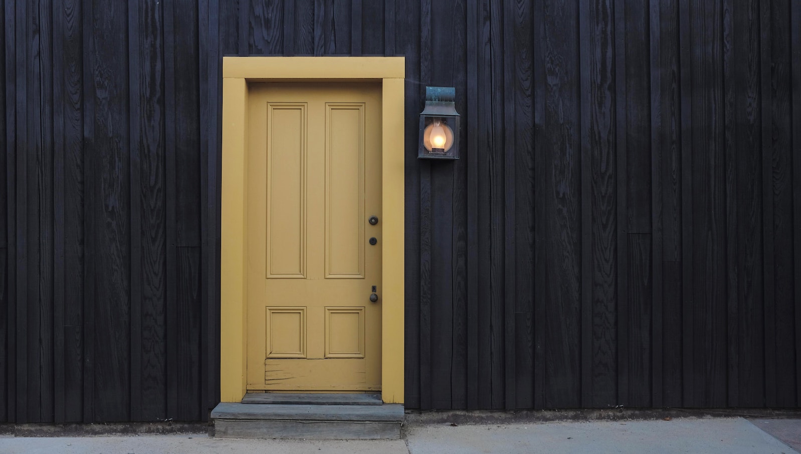 A yellow door against a black wooden wall with a light next to the door