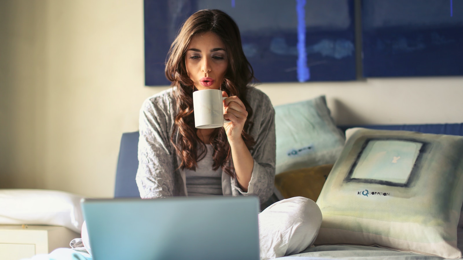 Woman on computer sitting in bed, sipping coffee