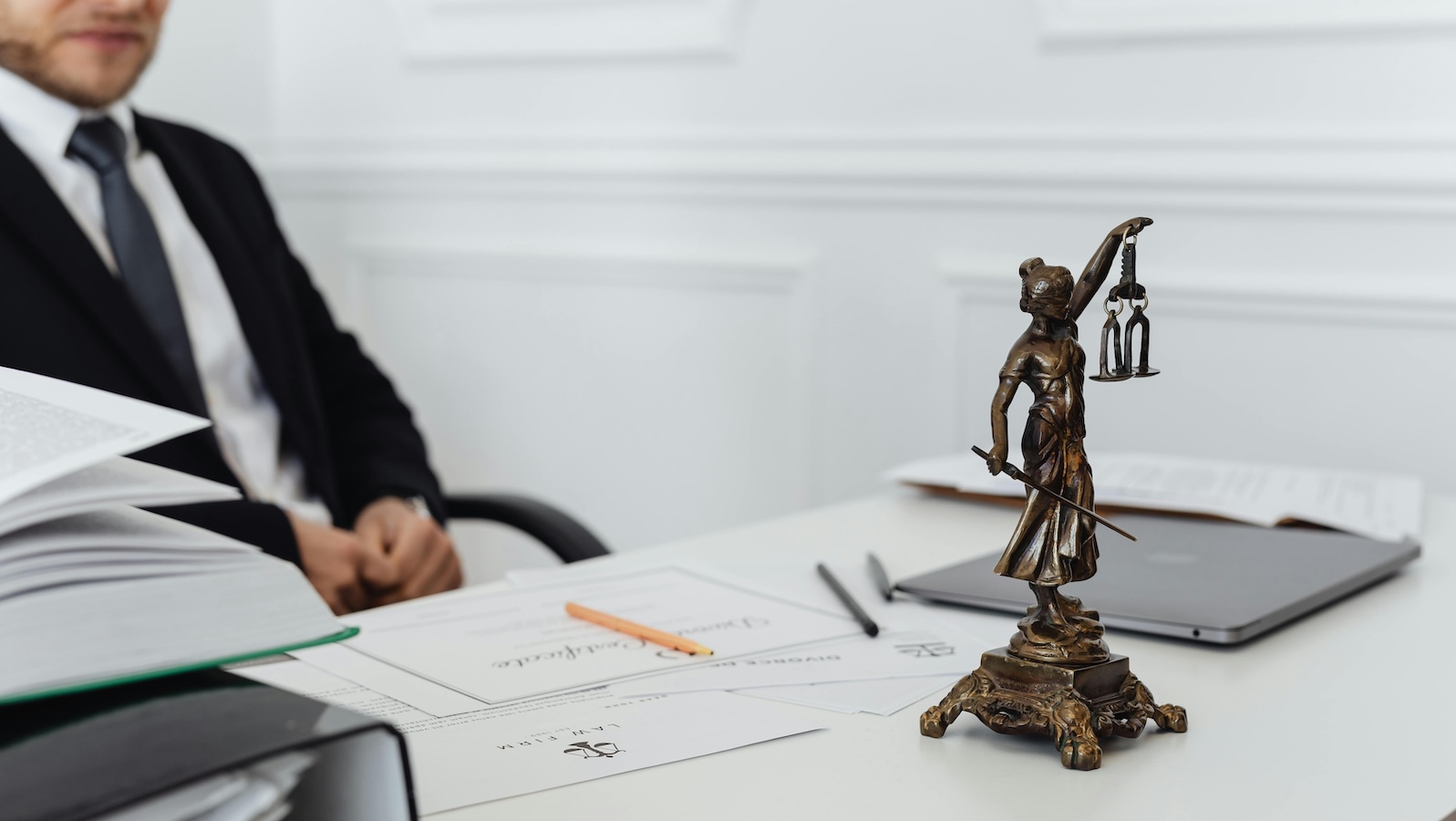 Man in a suit behind a white desk with papers on the desk and a statue for the scales of justice on the desk