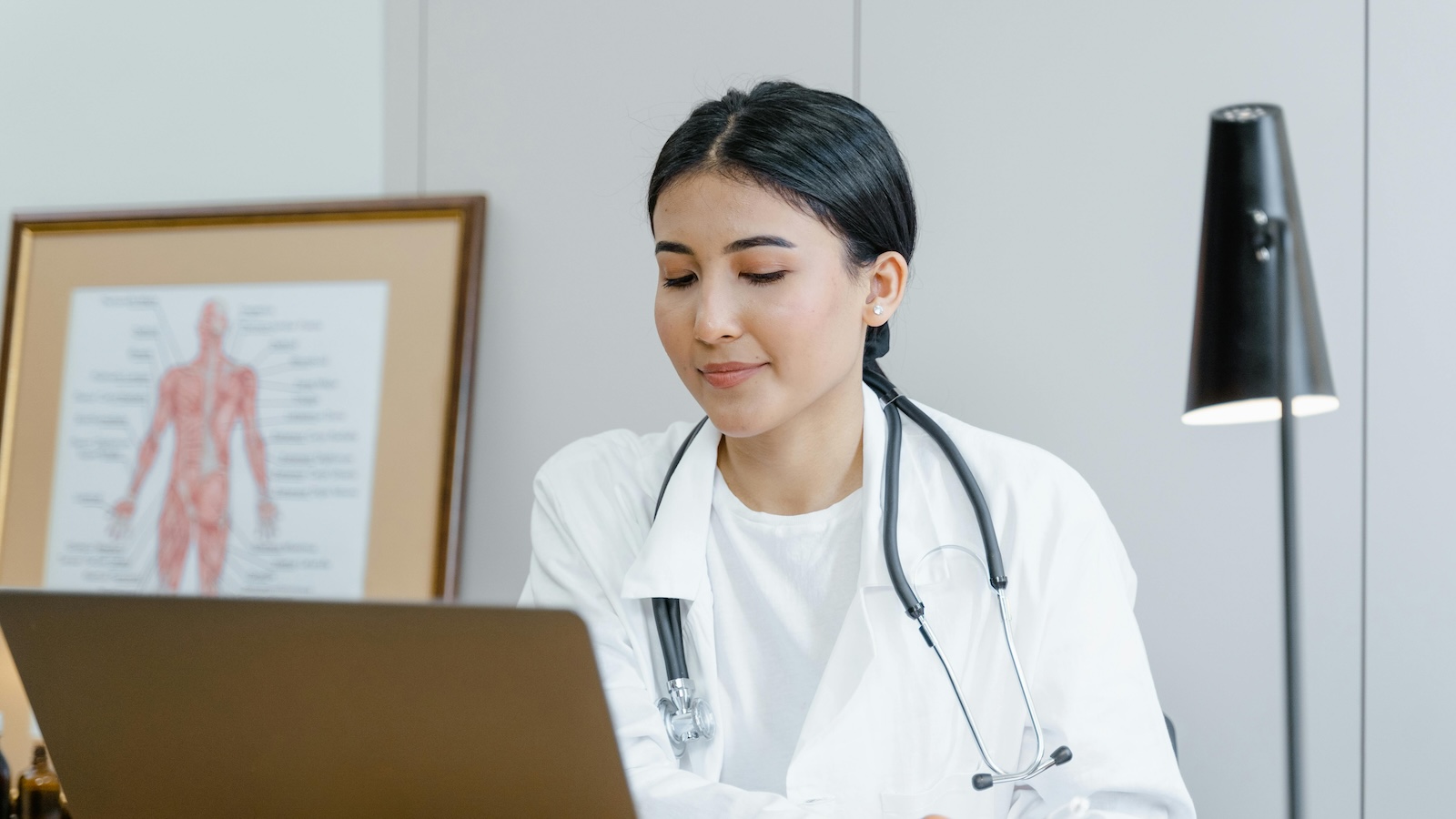 Dctor in a white coat with a stethoscope around her neck looking at a screen against a white office background