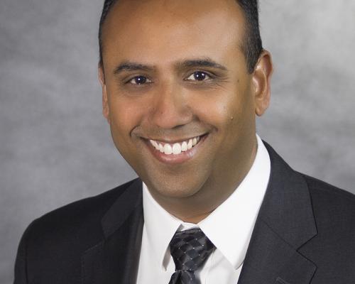 a photo of Murali Natarajan. The background is a textured greyscale and he is wearing a white button down, black suit jacket and black patterned tie