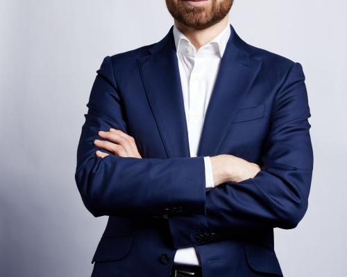 a photo of Henri de Combles de Nayves standing against a grey background in a navy blue suit and a white button down shirt 