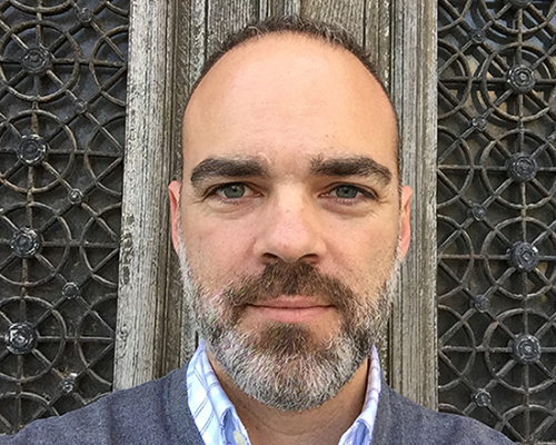 a headshot of Spiro Skias he is wearing a grey sweater over a blue and white plaid shirt and standing in front of a grey door