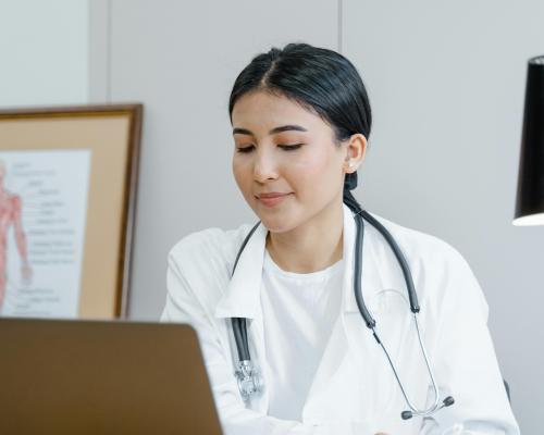 Dctor in a white coat with a stethoscope around her neck looking at a screen against a white office background