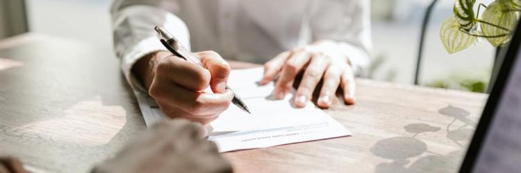 A person's hands writing an insurance forum. There is a laptop that is off to the side and a plant whose shadow is reflected on the table. 