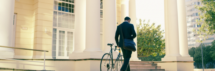 a man turned away from the camera, walking towards a white building. The man is in a suit and holding a bike