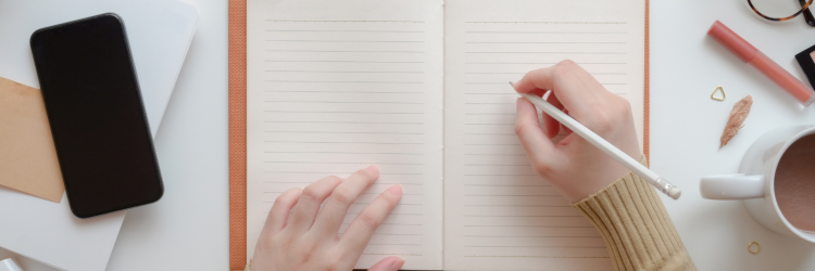 two hands writing in the notebook on a desk there is also a smartphone, a cup of coffee. two notebooks, a pen and a pair of glasses on the desk.