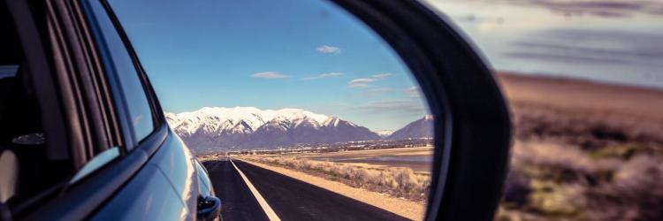 Close-up image of a car's side mirror showing a reflection of the road and mountains and blue sky behind it alongside an image in the foreground