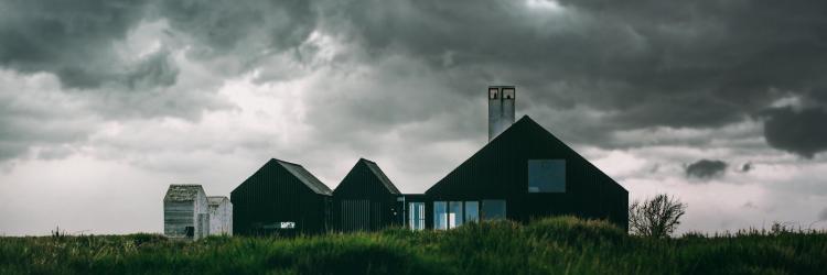A simple brown house in the countryside atop of tall green grass and beneath a dark and cloudy sky