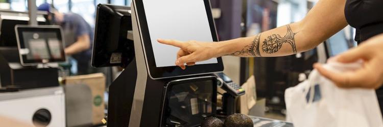 A woman touching the screen at a self checkout stand in a store with two avocados sitting on the scale